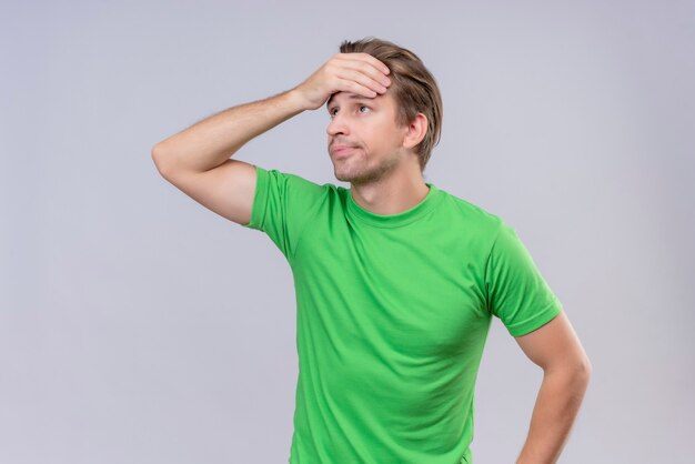 Jonge knappe man met een groen t-shirt opzij kijken met de hand op het hoofd voor een fout die over de witte muur staat 2