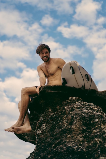 Jonge knappe man met een surfplank op een rots in de buurt van de oceaan.