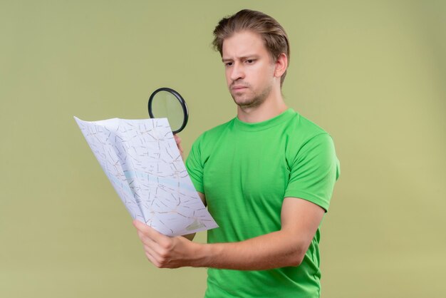 Jonge knappe man met groen t-shirt kaart kijken door vergrootglas met ernstige uitdrukking op gezicht staande over groene muur