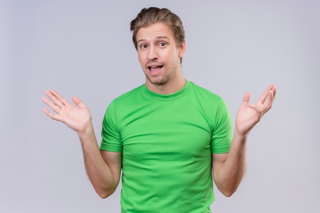 Jonge knappe man met groen t-shirt op zoek verward glimlachend spreidende armen