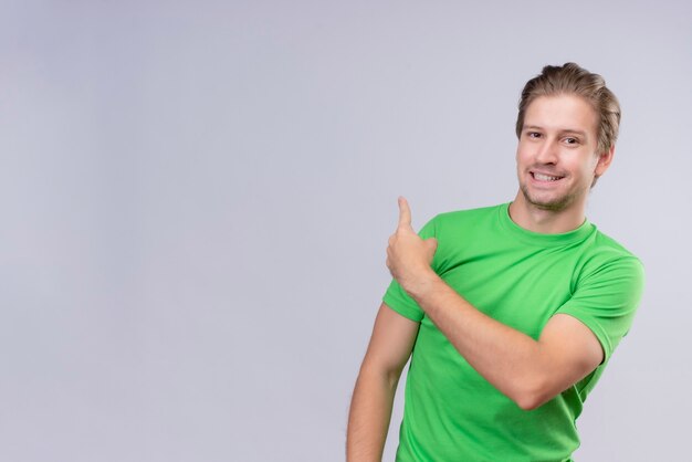 Jonge knappe man met groene t-shirt glimlachend vrolijk wijzend met de vinger naar iets achter staande over witte muur