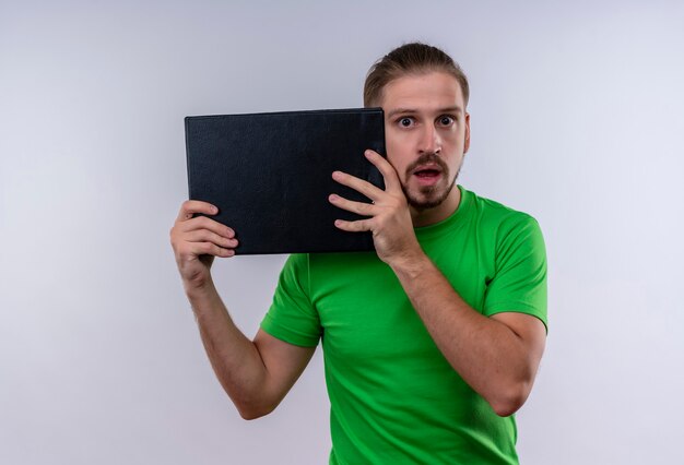 Jonge knappe man met groene t-shirt houden klembord kijken camera verrast staande op witte achtergrond