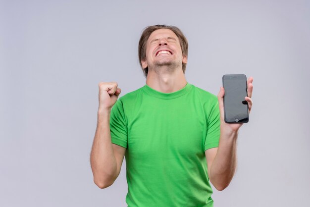 Jonge knappe man met groene t-shirt met mobiele telefoon gekke blije gebalde vuist verheugend zijn succes staande over witte muur