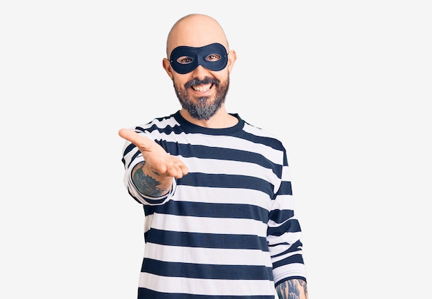 Gratis foto jonge knappe man met inbrekersmasker die vrolijk glimlacht en palmhand aanbiedt die hulp en acceptatie geeft.