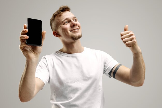 Jonge knappe man toont smartphonescherm geïsoleerd op grijze muur in shock met een verrassingsgezicht