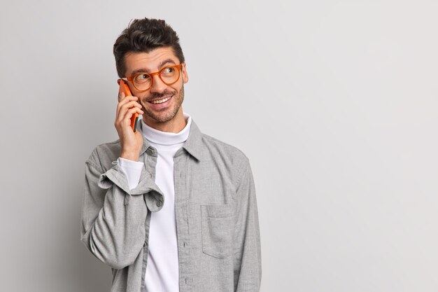 Jonge knappe mannelijke freelancer praat via mobiele telefoon heeft vrolijke uitdrukking geniet van mobiele tarieven en verbinding