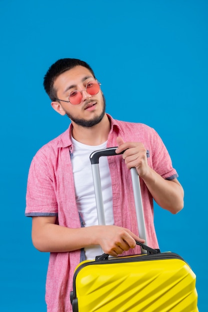 Jonge knappe reiziger kerel die zonnebril draagt die reiskoffer houdt die opzij kijkt met zelfverzekerde en ernstige uitdrukking op gezicht dat zich over blauwe achtergrond bevindt