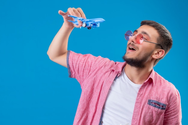 Jonge knappe reiziger kerel die zonnebril houdt die speelgoedvliegtuig speelt met hem op zoek gelukkig en positief glimlachend vrolijk staande over blauwe achtergrond
