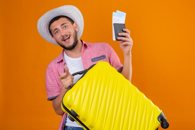 Jonge knappe reiziger kerel in zomer hoed bedrijf koffer en vliegtickets kijken camera verlaten en gelukkig lachend vrolijk klaar om te reizen staande over oranje achtergrond