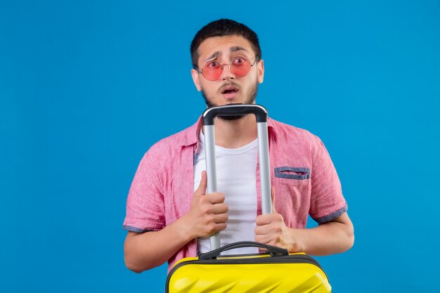 Jonge knappe reiziger man draagt een zonnebril bedrijf koffer kijken camera met verwarren uitdrukking op gezicht staande over blauwe achtergrond