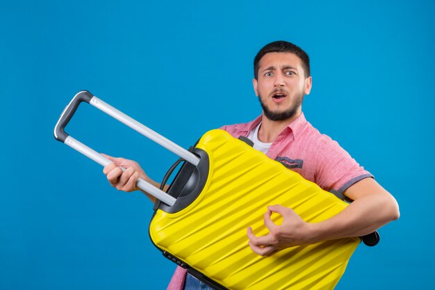 Jonge knappe reiziger man met koffer gebruikt als een gitaar kijken vrolijk met blij gezicht staande over blauwe achtergrond