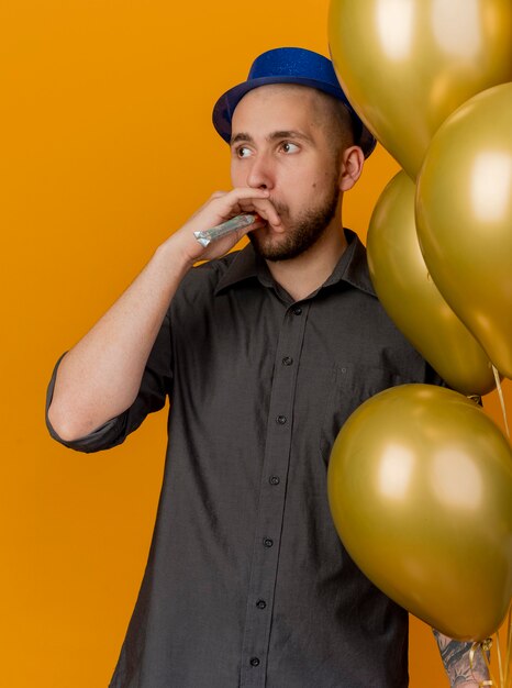 Jonge knappe Slavische partijkerel die partijhoed draagt die ballons houdt die partijventilator blaast die kant bekijkt die op oranje achtergrond wordt geïsoleerd