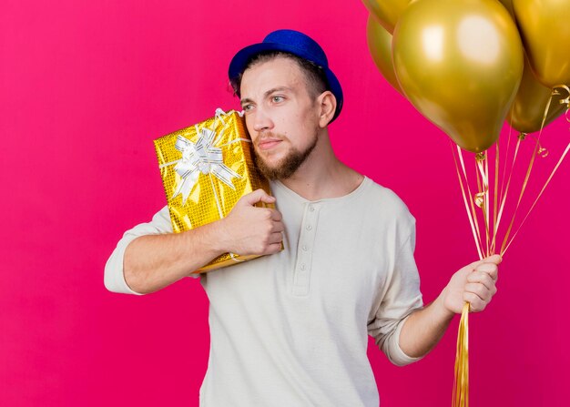 Jonge knappe Slavische partijkerel die partijhoed draagt die ballons houdt en gezicht met giftdoos aanraakt die kant bekijkt die op roze muur wordt geïsoleerd