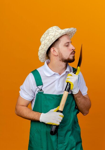 Jonge knappe slavische tuinman in uniform met hoed en tuinhandschoenen houden en kijken naar spade kus gebaar geïsoleerd op oranje muur