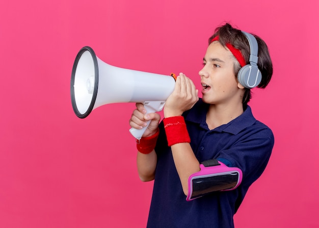 Jonge knappe sportieve jongen die hoofdband en polsbandjes en koptelefoon draagt, telefoonarmband met beugels, kijkt rechtuit pratend door spreker geïsoleerd op karmozijnrode muur
