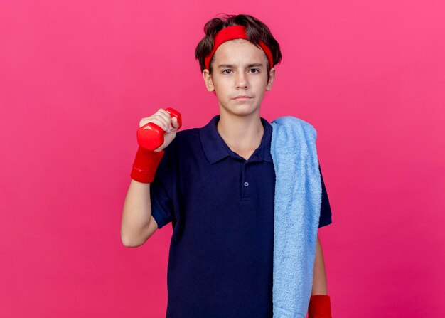 Jonge knappe sportieve jongen die hoofdband en polsbandjes met beugels en handdoek op schouder draagt ?? die aan de voorzijde houdt die halter houdt die op roze muur met exemplaarruimte wordt geïsoleerd