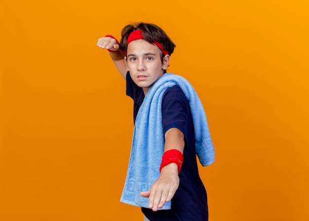 Jonge knappe sportieve jongen die hoofdband en polsbandjes met beugels en handdoek op schouder draagt ?? die zich in profiel bekijken die op oranje achtergrond met exemplaarruimte wordt geïsoleerd