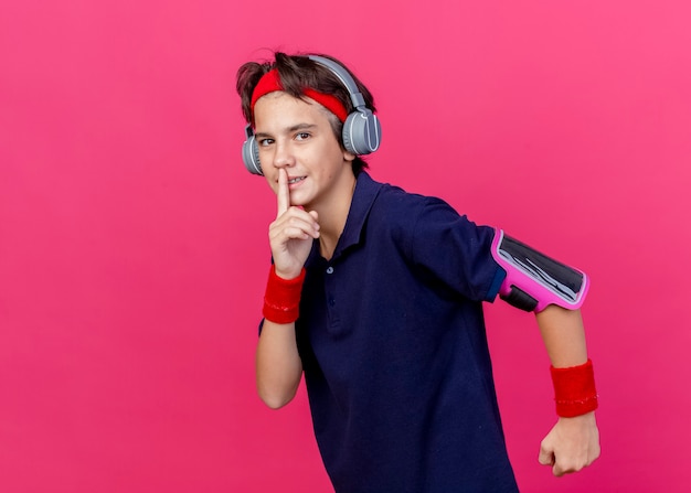 Jonge knappe sportieve jongen met hoofdband en polsbandjes en koptelefoon telefoon armband met beugels kijken camera uitgevoerd gebaren stilte geïsoleerd op karmozijnrode achtergrond met kopie ruimte