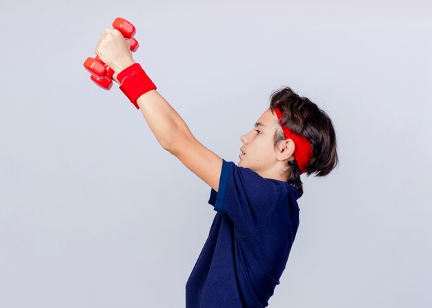 Jonge knappe sportieve jongen met hoofdband en polsbandjes met beugels staan ?? in profiel te bekijken op zoek recht omhoog halters geïsoleerd op een witte achtergrond met kopie ruimte