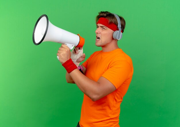 Jonge knappe sportieve man met hoofdband en polsbandjes en koptelefoon en telefoonarmband met gewonde pols omwikkeld met verband in profielweergave pratend door spreker