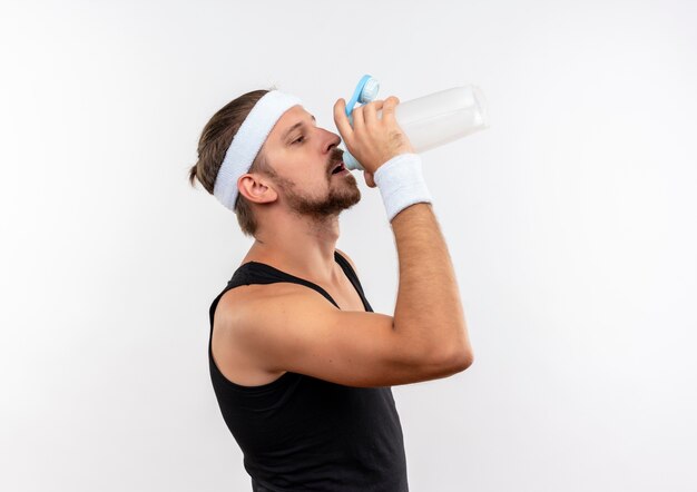 Jonge knappe sportieve man met hoofdband en polsbandjes permanent in profiel te bekijken en drinkwater uit fles geïsoleerd op witte ruimte