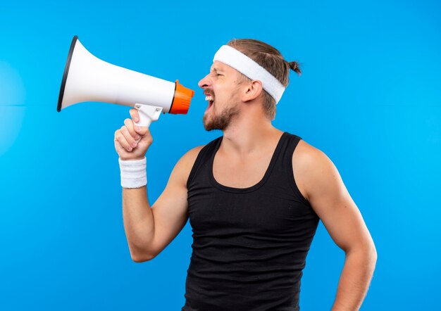 Jonge knappe sportieve man met hoofdband en polsbandjes schreeuwen in luide luidspreker geïsoleerd op blauwe ruimte