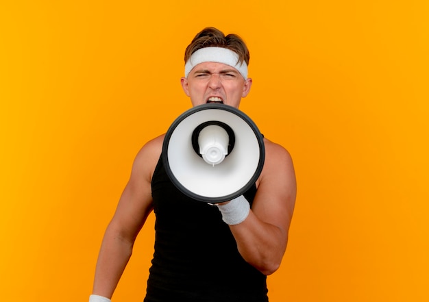 Jonge knappe sportieve man met hoofdband en polsbandjes schreeuwen in luide luidspreker op camera geïsoleerd op een oranje achtergrond met kopie ruimte