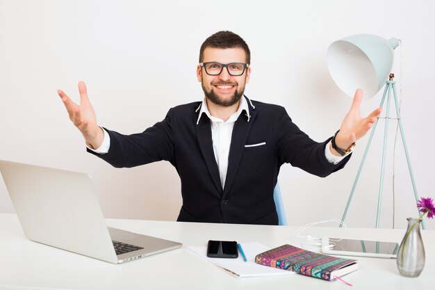 Jonge knappe stijlvolle hipster man in zwarte jas aan kantoor tafel, zakelijke stijl, wit overhemd, geïsoleerd, bezig met laptop, opstarten, werkplek, open armen hallo groet, glimlachen, blij, positief