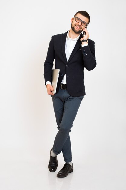 Jonge knappe stijlvolle hipster man in zwarte jas, zakelijke stijl, wit overhemd, geïsoleerd, witte achtergrond, glimlachen, staande volledige hoogte, op zoek zelfverzekerd, met laptop, praten over smartphone