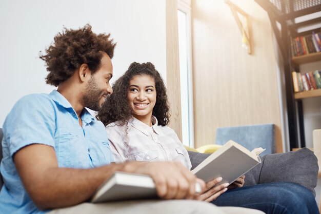 Jonge knappe student twee in vrijetijdskleding die op bank in heldere moderne bibliotheek na studie zitten, glimlachen, spreken, boeken lezen, informatie zoeken voor het project van het graduatieteam.