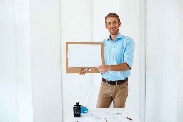 Gratis foto jonge knappe vrolijke glimlachende zakenman die zich bij lijst bevinden die houten klembord met wit blad houden. licht modern kantoorinterieur