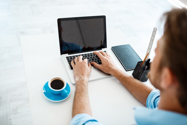 Jonge knappe vrolijke zekere peinzende zakenmanzitting bij lijst die aan laptop met opzij kop van koffie werken. Witte moderne kantoor interieur