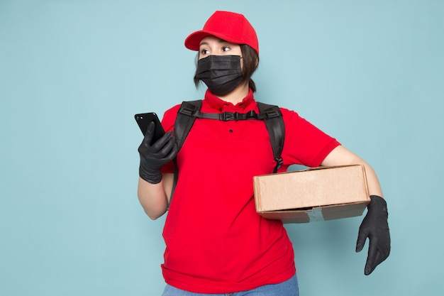 jonge koerier in rode polo rode dop zwart steriel beschermend masker zwarte rugzak met pakket met behulp van de telefoon op blauw