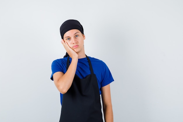 Jonge kok man in t-shirt, schort leunend op de hand en er attent uitziend, vooraanzicht.