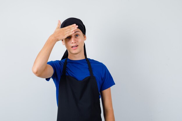 Jonge kok man in t-shirt, schort met hand op voorhoofd en vergeetachtig, vooraanzicht.