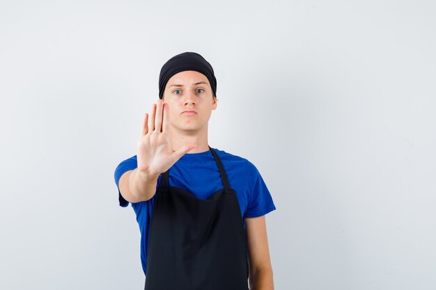 Jonge kok man in t-shirt, schort met stopgebaar en serieus, vooraanzicht.