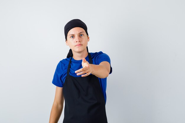 Jonge kok wijst naar voren in t-shirt, schort en ziet er somber uit. vooraanzicht.