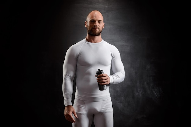 Gratis foto jonge krachtige sportman in witte kleding glimlachend, kijkend naar voren, fles over zwarte muur houdend