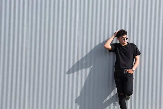Jonge krullende man in zonnebril en zwarte kleding leunend op grijze muur