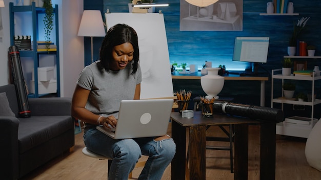 Jonge kunstenaar van afro-amerikaanse etniciteit met behulp van laptopcomputer op zoek naar inspiratie voor tekentechniek in creativiteit kamer. zwarte vrouw met moderne technologie bezig met meesterwerk