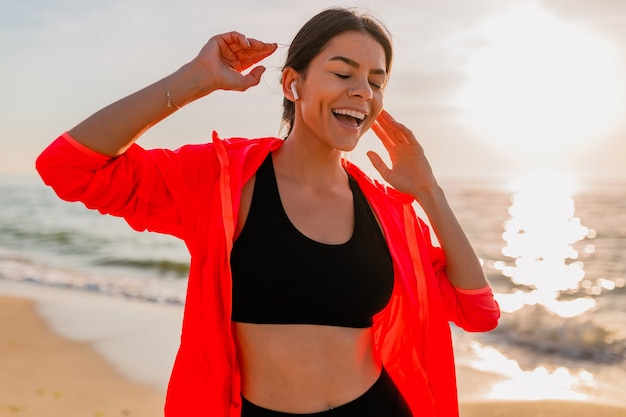 Jonge lachende aantrekkelijke slanke vrouw doet sport in de ochtend zonsopgang dansen op zee strand in sportkleding, gezonde levensstijl, luisteren naar muziek op oortelefoons, roze windjack dragen, plezier maken