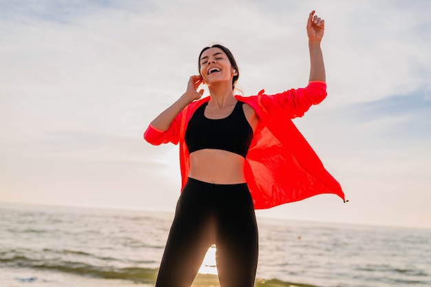 Jonge lachende aantrekkelijke slanke vrouw doet sport in de ochtend zonsopgang dansen op zee strand in sportkleding, gezonde levensstijl, luisteren naar muziek op oortelefoons, roze windjack dragen, plezier maken