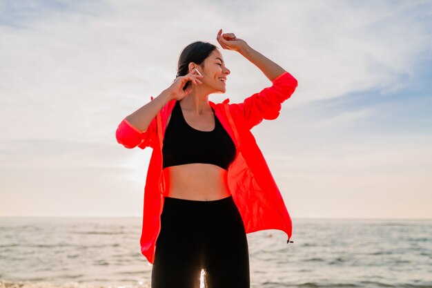 Jonge lachende aantrekkelijke slanke vrouw doet sport in de ochtend zonsopgang dansen op zee strand in sportkleding, gezonde levensstijl, luisteren naar muziek op oortelefoons, roze windjack dragen, plezier maken