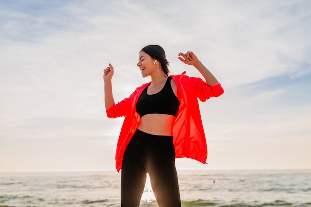 Jonge lachende aantrekkelijke slanke vrouw doet sport in de ochtend zonsopgang dansen op zee strand in sportkleding, gezonde levensstijl, luisteren naar muziek op oortelefoons, roze windjack dragen, plezier maken