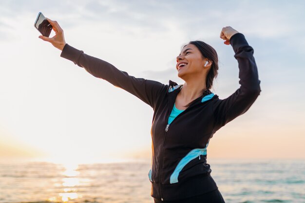 Jonge lachende aantrekkelijke slanke vrouw sport oefeningen doen op ochtend zonsopgang strand in sportkleding, gezonde levensstijl, luisteren naar muziek op oortelefoons, selfie foto op telefoon er sterk uitzien