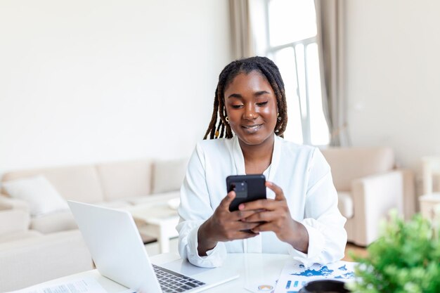 Jonge lachende Afrikaanse zakenvrouw met behulp van smartphone in de buurt van computer op kantoor