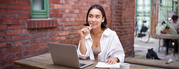 Jonge lachende aziatische vrouw met draadloze oortelefoons die in coworking kantoorruimte zitten met laptop
