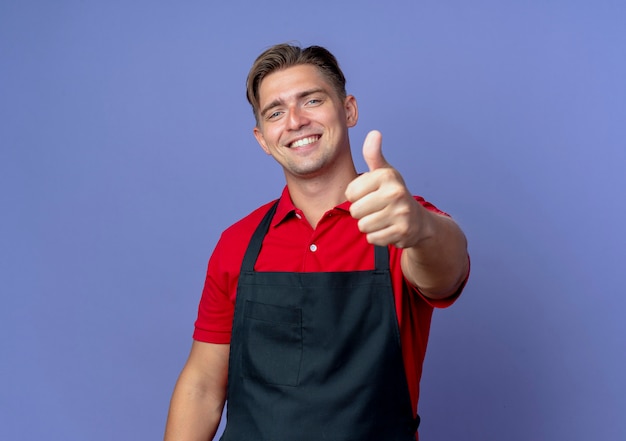 Jonge lachende blonde mannelijke kapper in uniforme duimen omhoog geïsoleerd op violette ruimte met kopie ruimte