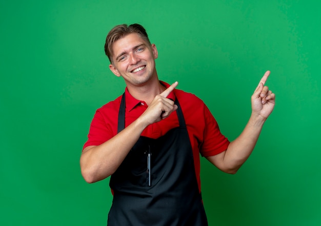 Jonge lachende blonde mannelijke kapper in uniforme punten aan de zijkant met twee handen geïsoleerd op groene ruimte met kopie ruimte