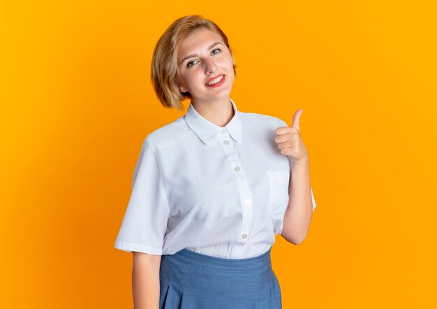 Jonge lachende blonde Russische meisje duimen omhoog geïsoleerd op een oranje achtergrond met kopie ruimte